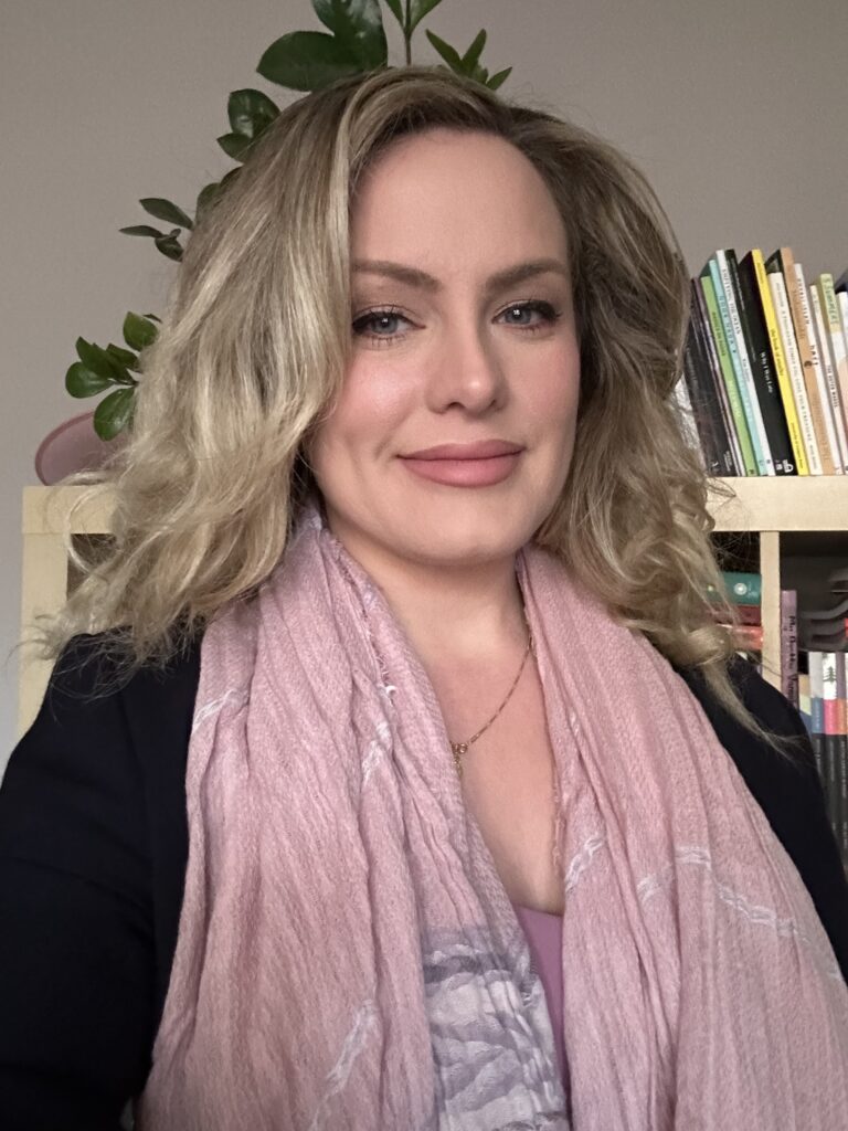 Headshot of Margo LaPierre, a white woman in her 30s with long wavy blonde hair, wearing a pink scarf, pink lipstick, and a navy blazer smiling at the camera. Behind her is a shelf of poetry books and a ZZ plant.