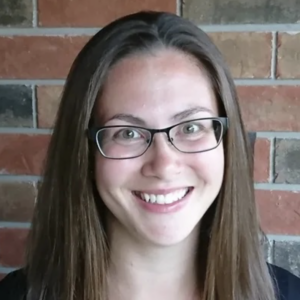 Headshot of Amanda Feeney. Amanda has long brown hair and light skin and is wearing dark-rimmed glasses. She is standing against a brick wall.
