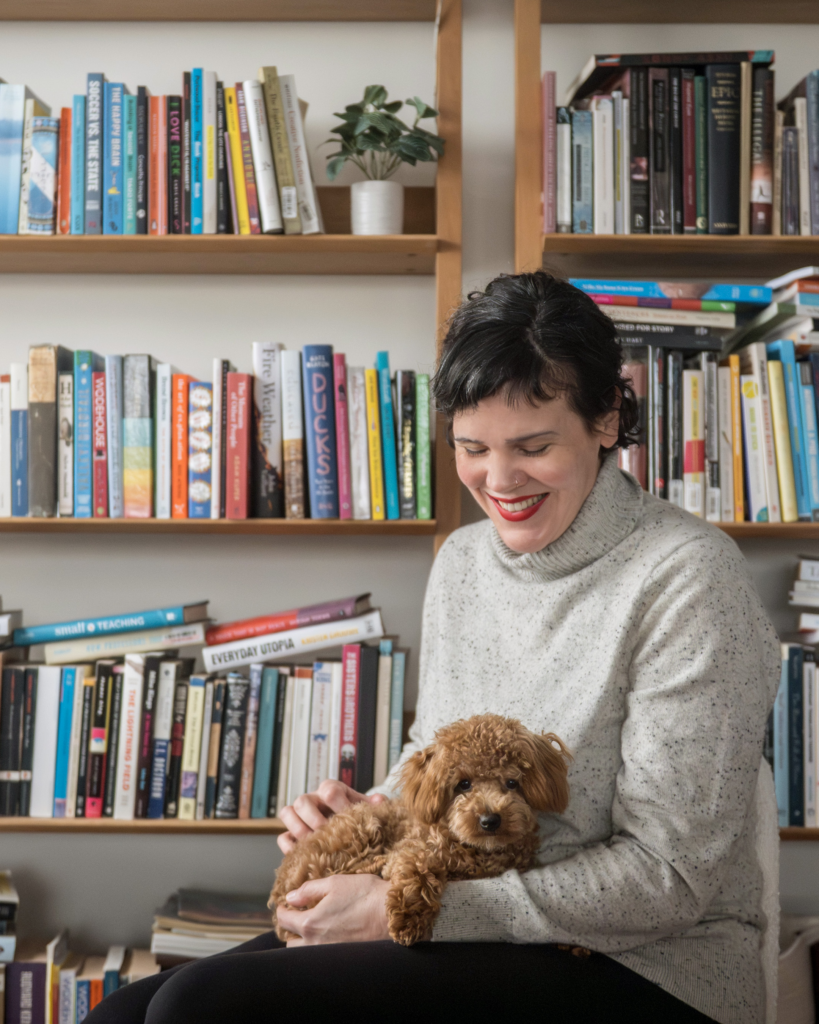 A white lady with black hair and red lipstick holds a small brown puppy on her lap. Behind her is a messy bookcase stacked with books.