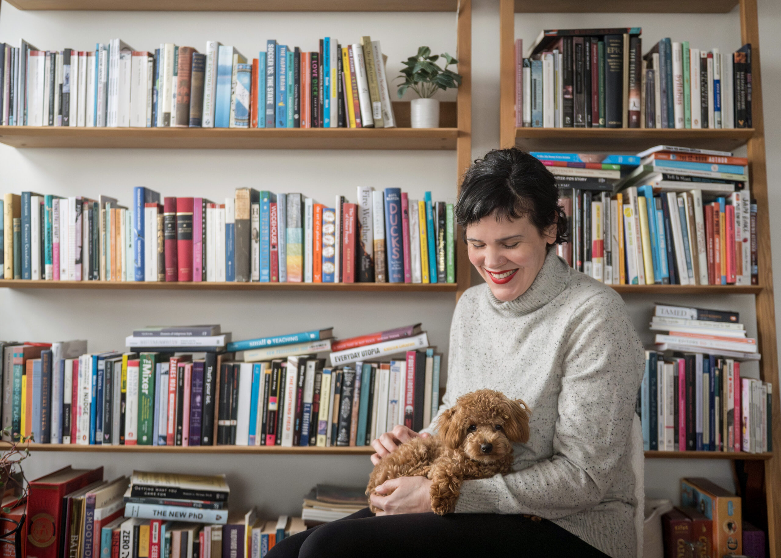 A white lady with black hair and red lipstick holds a small brown puppy on her lap. Behind her is a messy bookcase stacked with books.
