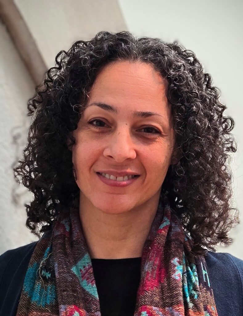 Leïta has dark, shoulder-length curly hair and brown eyes. She is smiling and is wearing a blue top with a multi-coloured scarf around her shoulders.