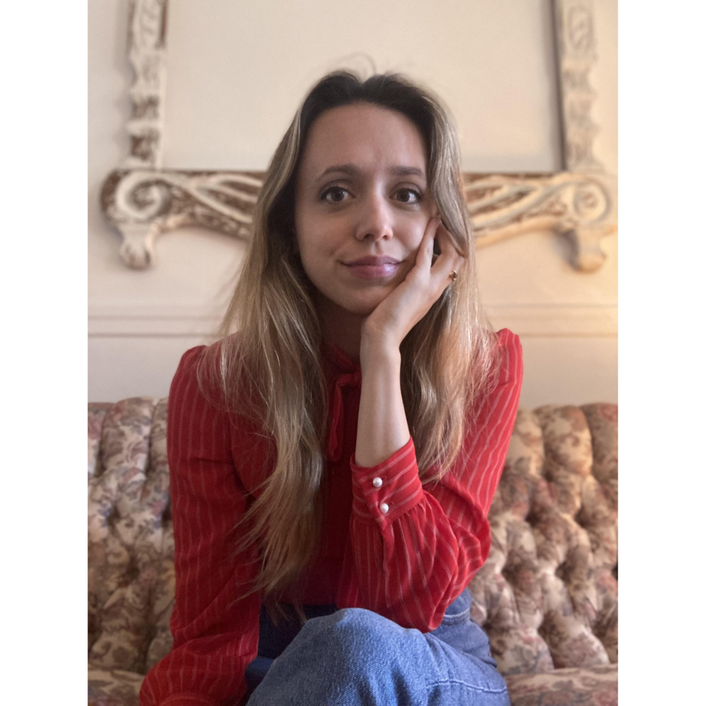 Headshot of Holly Vestad. She has long blond hair and fair skin and is holding her left hand up to her face. She is wearing a striped red shirt.
